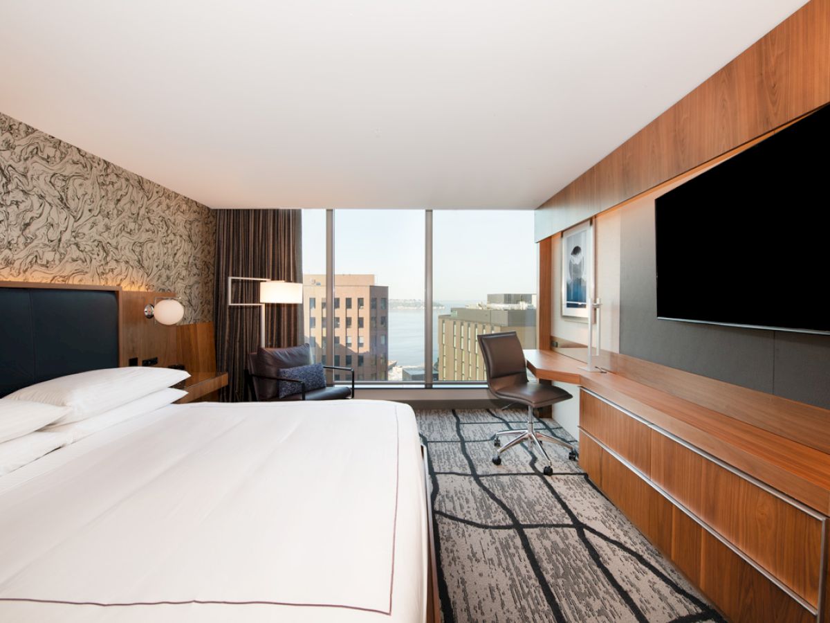 This image shows a modern hotel room with a large bed, a desk, a chair, and a flat-screen TV. A window provides a view of buildings and water.