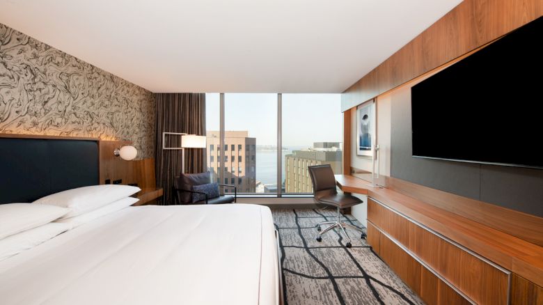 This image shows a modern hotel room with a large bed, a desk, a chair, and a flat-screen TV. A window provides a view of buildings and water.