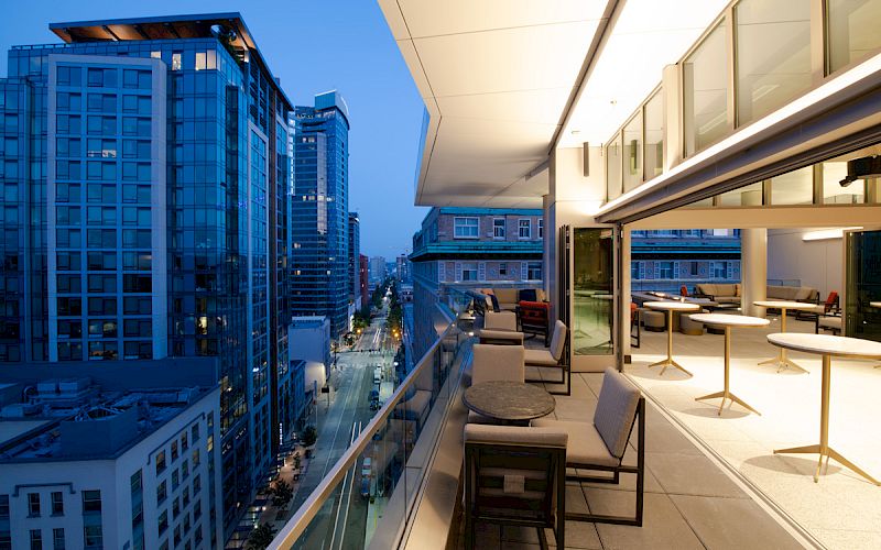 A modern balcony overlooking a cityscape with tall buildings, tables, chairs, and a cozy setup, all illuminated during dusk time.