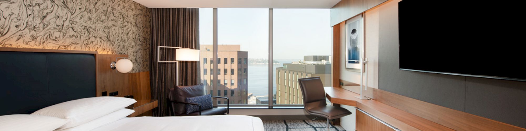 A modern hotel room features a large bed, desk, chair, wall-mounted TV, city view through large windows, and contemporary decor with wooden accents.