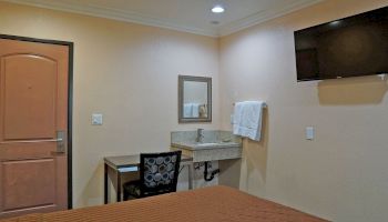 The image shows a simple hotel room with a bed, a wall-mounted TV, a small desk and chair, a sink with a mirror, and a towel on a rack.