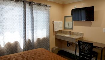 A small room with a bed, patterned curtains, a wall-mounted TV, a sink with mirror, a towel hanging, a trash can, and a desk with a chair.