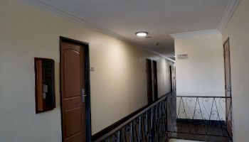 This image shows a hallway with several doors, a railing, a light fixture, and a sign on the wall. The walls are painted light yellow.