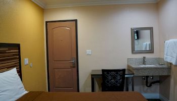 A hotel room features a bed, a desk with a chair, a sink with a mirror, and a door. The walls are painted orange and beige, with a towel hanging.