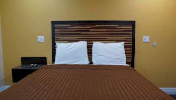 A neatly made bed with a brown blanket, two white pillows, a modern headboard, and a small side table with a TV remote on it.