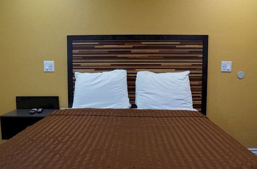 A neatly made bed with a brown blanket, two white pillows, a modern headboard, and a small side table with a TV remote on it.