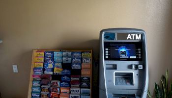 The image shows an ATM machine next to a display rack containing various brochures and informative pamphlets in a well-lit area.