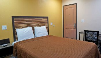 The image shows a small, neat bedroom with a brown bedspread, matching pillows, a headboard, a desk, a chair, and a closed door.