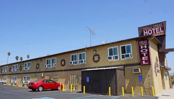 The image shows the exterior of the Holly Crest Hotel, a two-story building with a red car parked outside and signage visible on the structure.