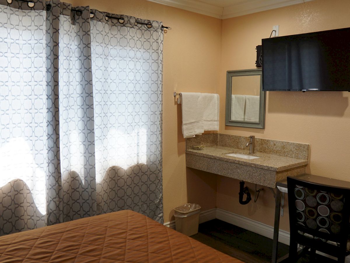 The image shows a hotel room with a bed, a small desk and chair, a wall-mounted TV, a sink area with a towel, and curtains covering a window.