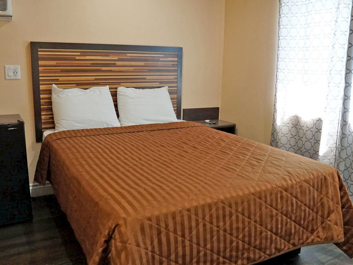 A small bedroom with a double bed, brown quilt, wooden headboard, pillows, mini-fridge, curtains with a pattern, and a wall-mounted air conditioner.