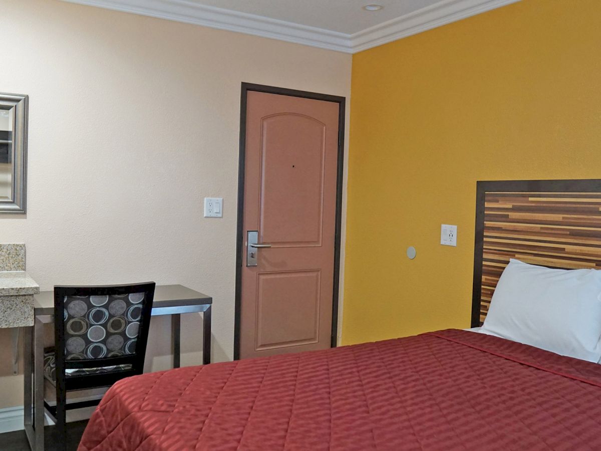 The image shows a hotel room with a bed, a desk with a chair, a sink and mirror, and a closed door with a beige and yellow wall.