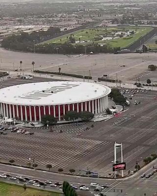 An aerial view of a large, circular arena with surrounding parking lots, sparse trees, adjacent roads, and some distant buildings in the background.