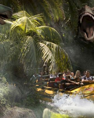 People on an amusement park water ride surrounded by animatronic dinosaurs and lush vegetation, creating a thrilling and adventurous atmosphere.